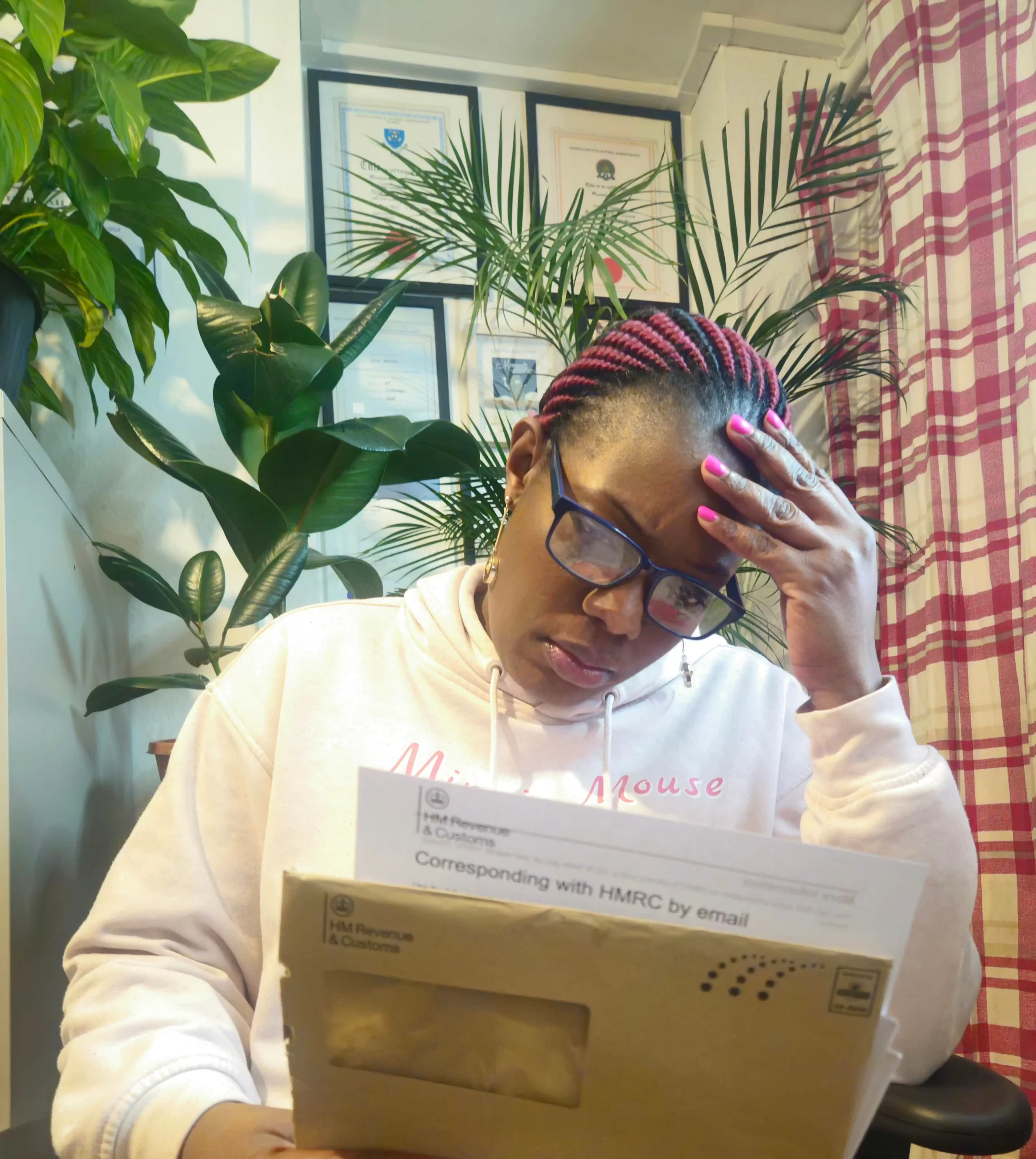 A woman reading a letter from HMRC, looking concerned, seated at her desk with framed certificates and plants in the background.