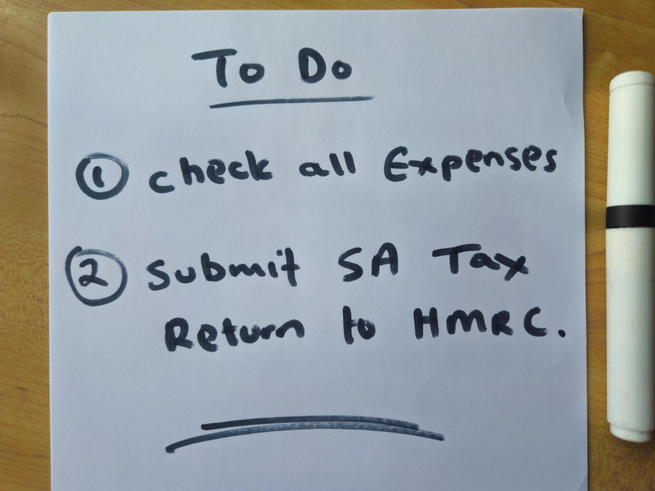 Handwritten to-do list with two items: “1. Check all expenses” and “2. Submit SA Tax Return to HMRC”, alongside a black marker on a wooden desk.
